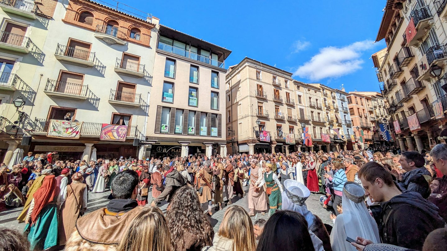 Persone in costume storico in una piazza soleggiata con striscioni.