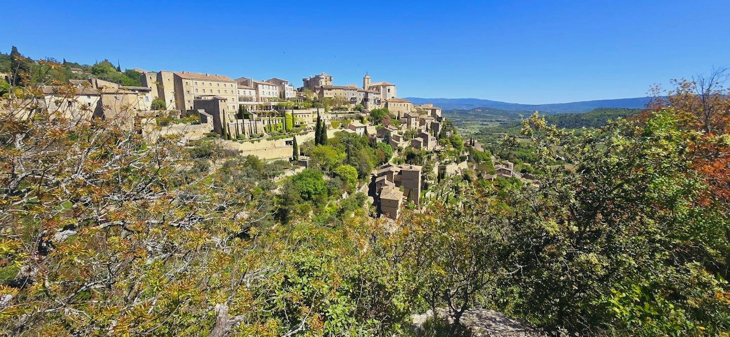 Villaggio soleggiato in pietra con tetti in terracotta su una collina verde.